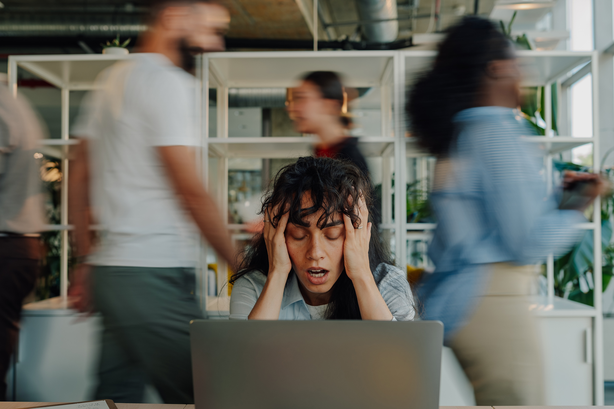 Eine Frau sitzt an einem Tisch vor einem Laptop und hält beide Hände an den Kopf.